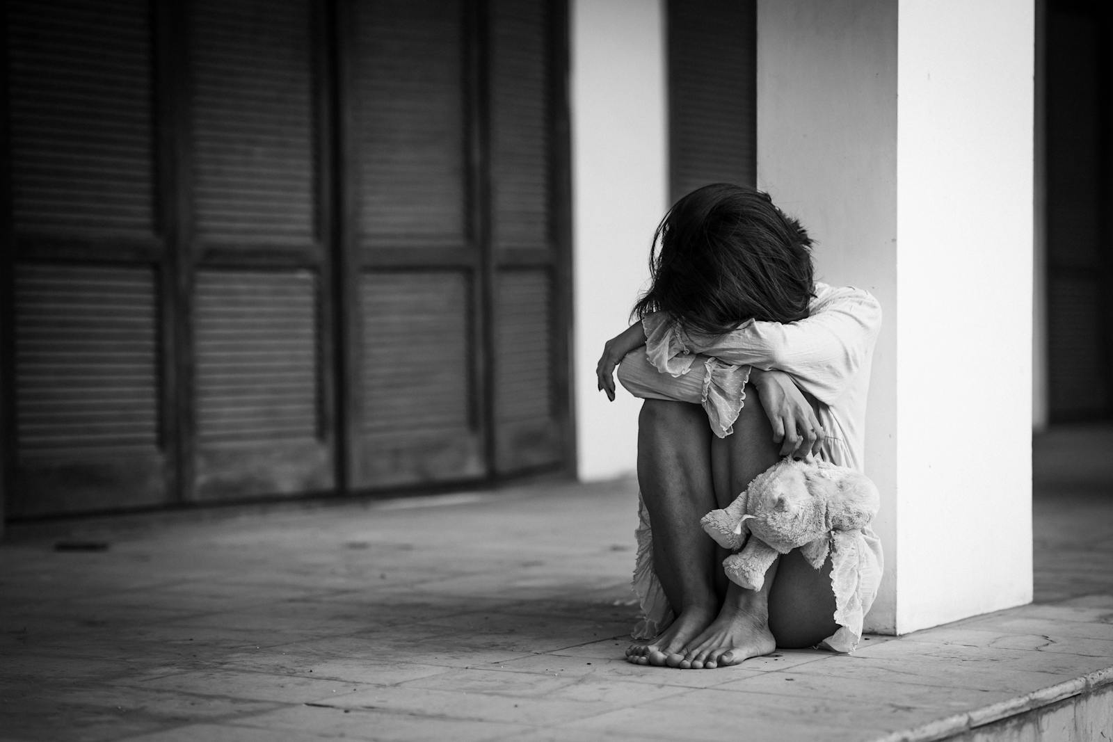 A dramatic black and white photograph of a woman sitting alone with a stuffed toy, evoking emotions of solitude and contemplation.